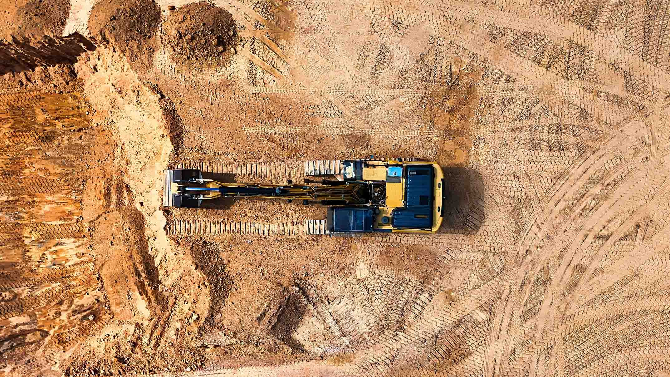 Blick von oben auf einen gelben Bagger, der auf einer großen Baustelle Erdarbeiten durchführt.