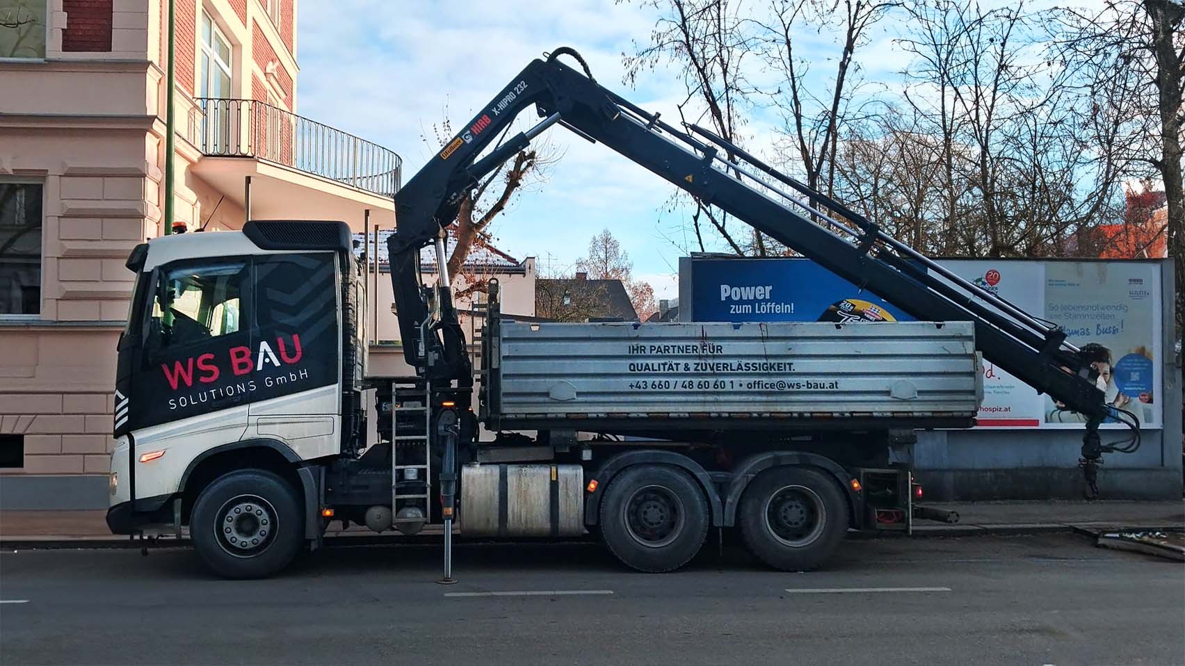 LKW mit Kranausleger und Aufschrift WS BAU Solutions GmbH, steht vor einem Haus an einer Straße.