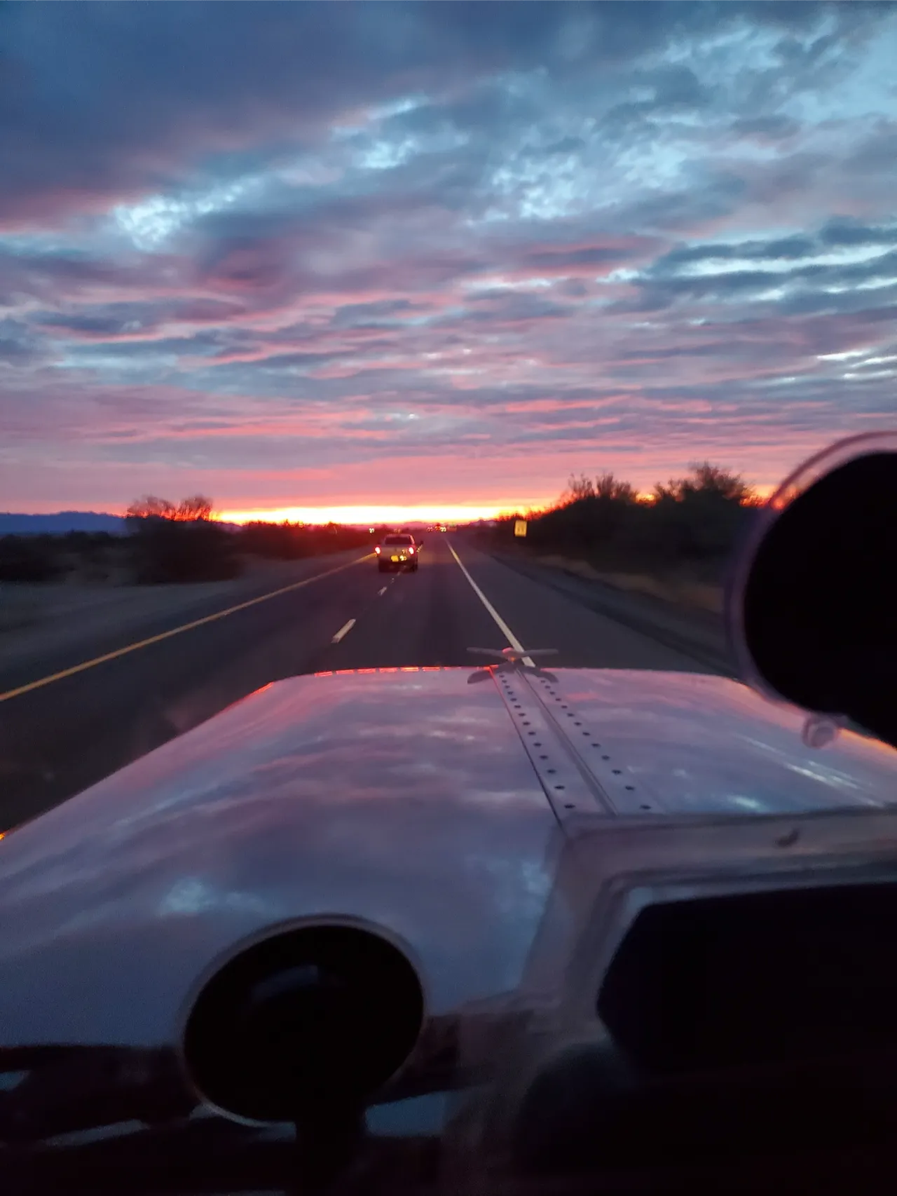 Heavy-duty truck transport heading through Fontana, CA at sunrise, highway lanes ahead with glowing sky reflected on the hood.