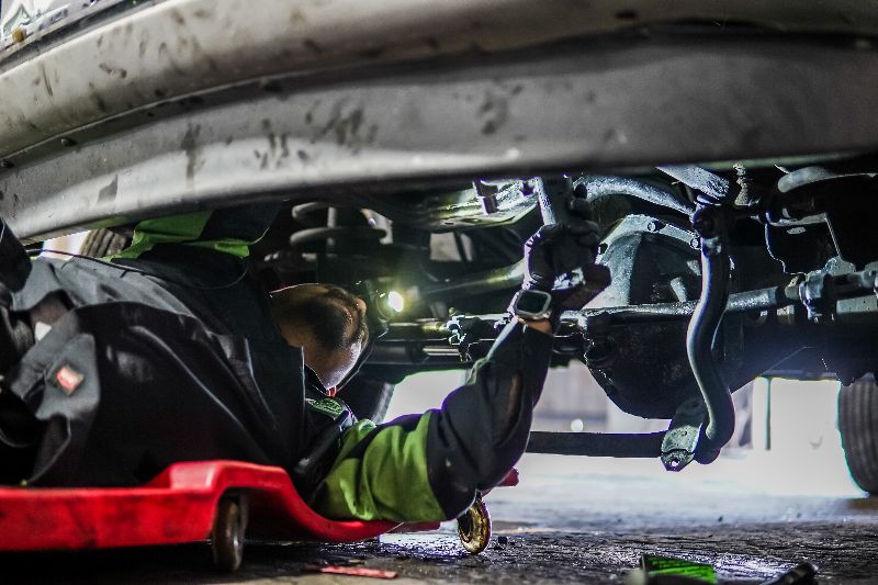 Mechanic inspecting heavy-duty truck undercarriage, checking suspension and steering components while lying on creeper in repair shop garage.