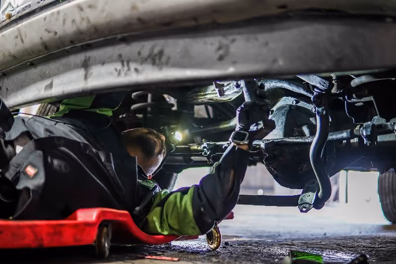 Mechanic inspecting heavy-duty truck undercarriage, checking suspension and steering components while lying on creeper in repair shop garage.