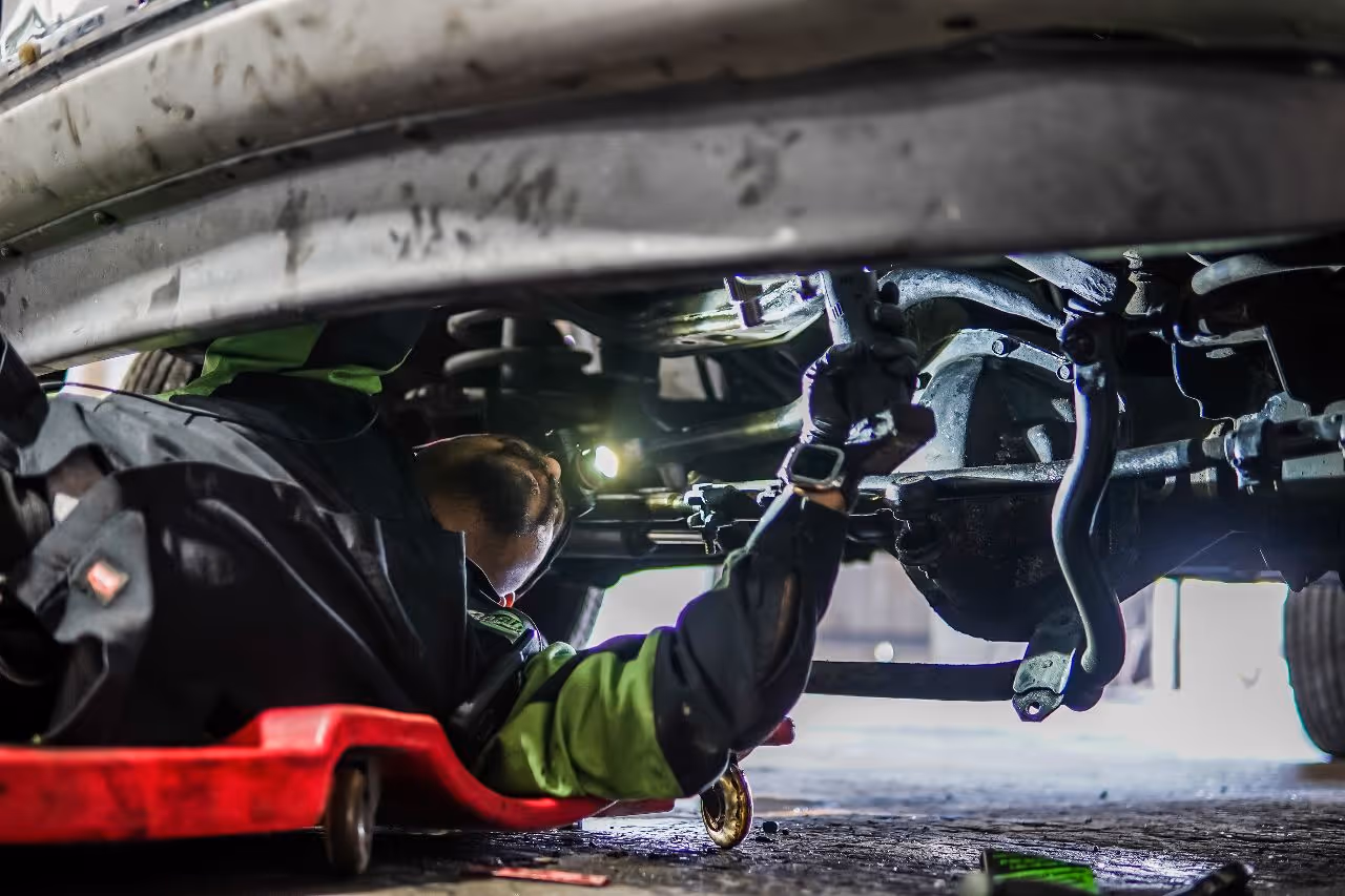Mechanic inspecting heavy-duty truck undercarriage, checking suspension and steering components while lying on creeper in repair shop garage.