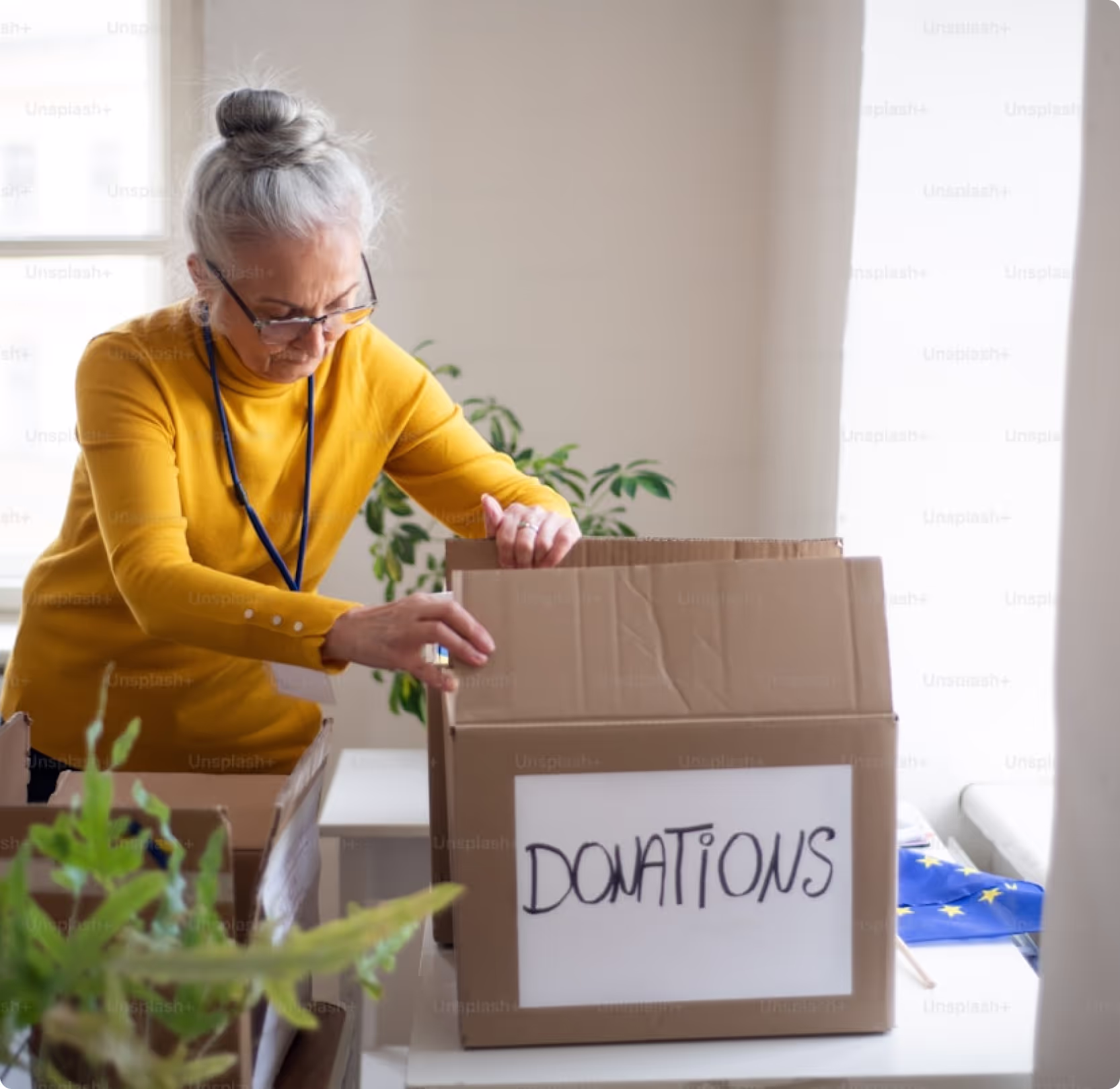 An older woman with gray hair pulled back in a bun and wearing glasses and a yellow sweater is packing items into a brown cardboard box labeled "DONATIONS" in black marker. 