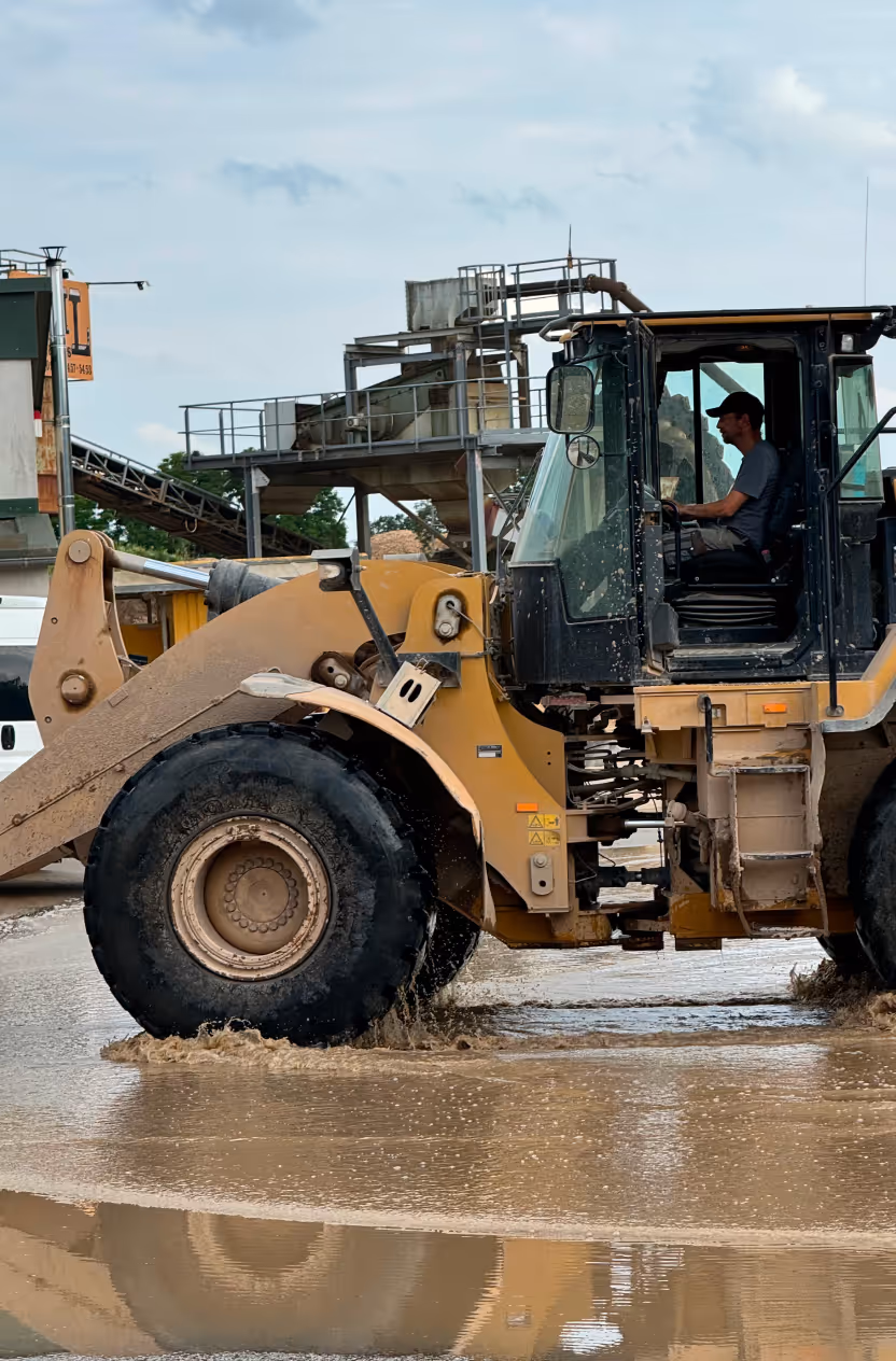 Baumaschine fährt durch eine schlammige Wasserpfütze auf einer Baustelle mit einem Fahrer im Führerhaus.