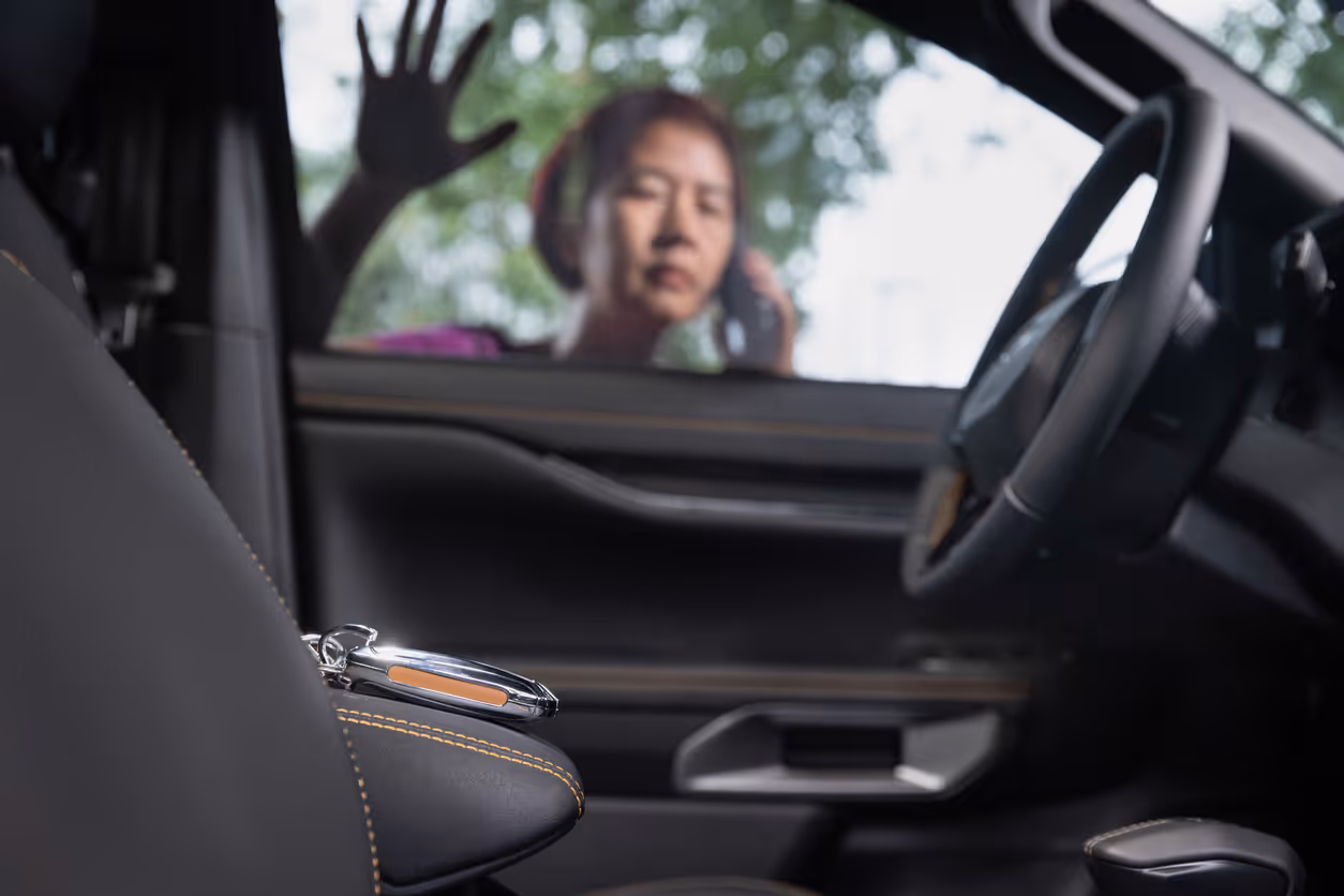 Car interior with car key on the armrest and a blurred person outside the window holding a phone and raising a hand.