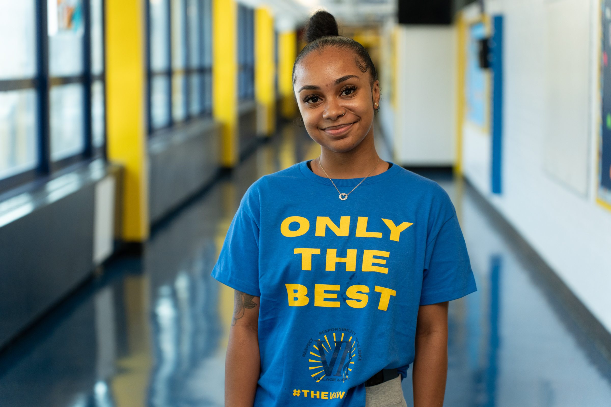 Ms K Arias smiling in school hallway wearing Only The Best shirt