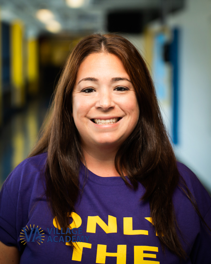 Ms. Meloni smiling in a Village Academy shirt in school hallway