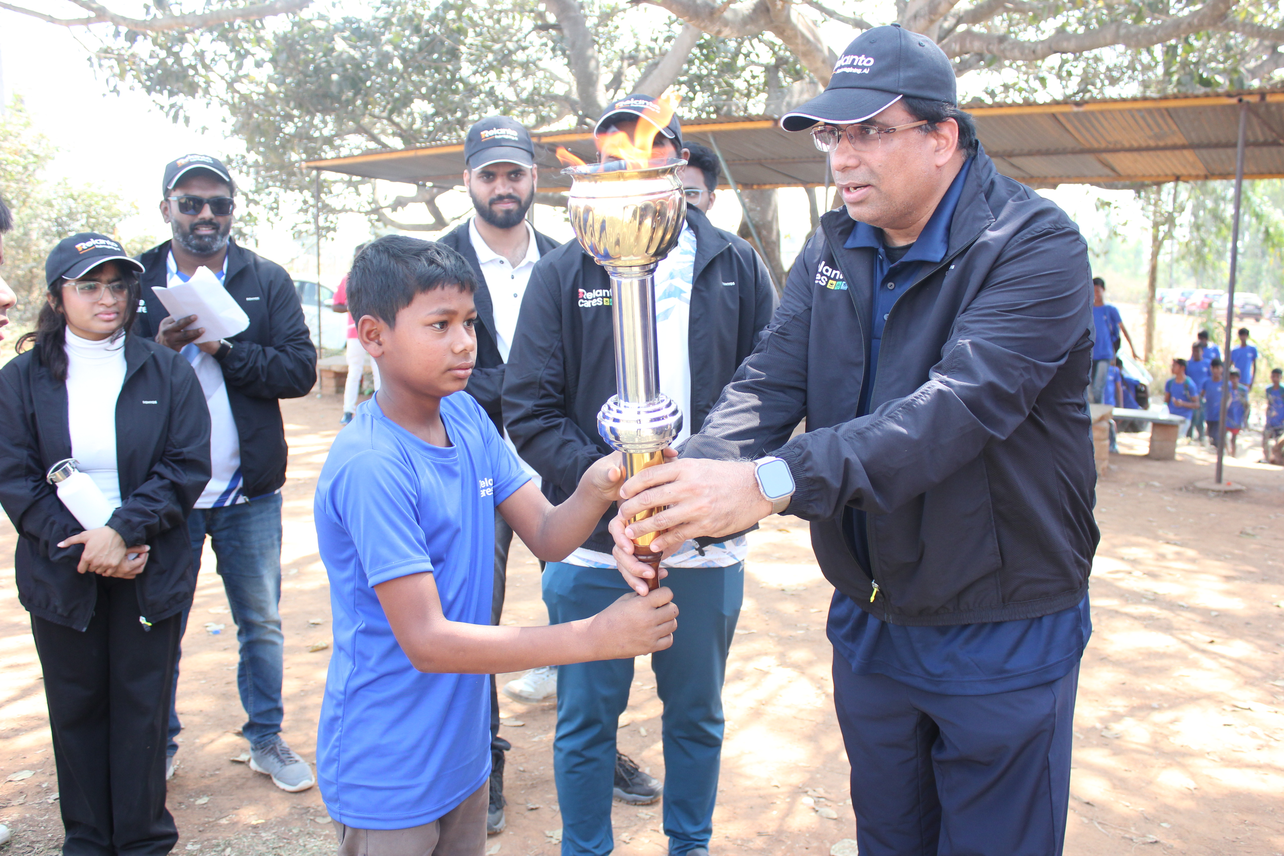 A boy hands a lit torch to a man in a black jacket, surrounded by people in a casual outdoor setting.