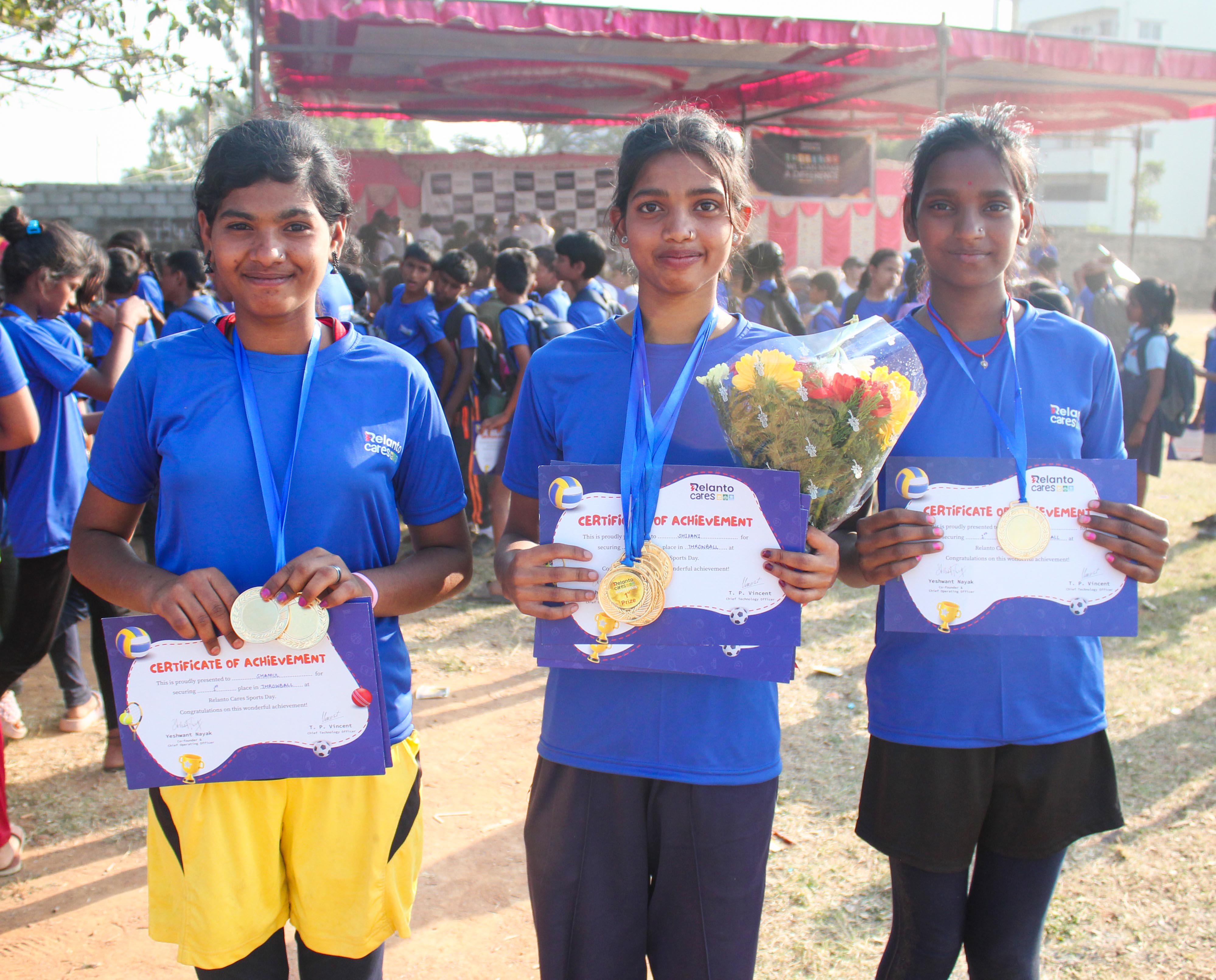 Three young girls in blue shirts proudly display medals, certificates, and a bouquet, celebrating their achievements at a sports event.