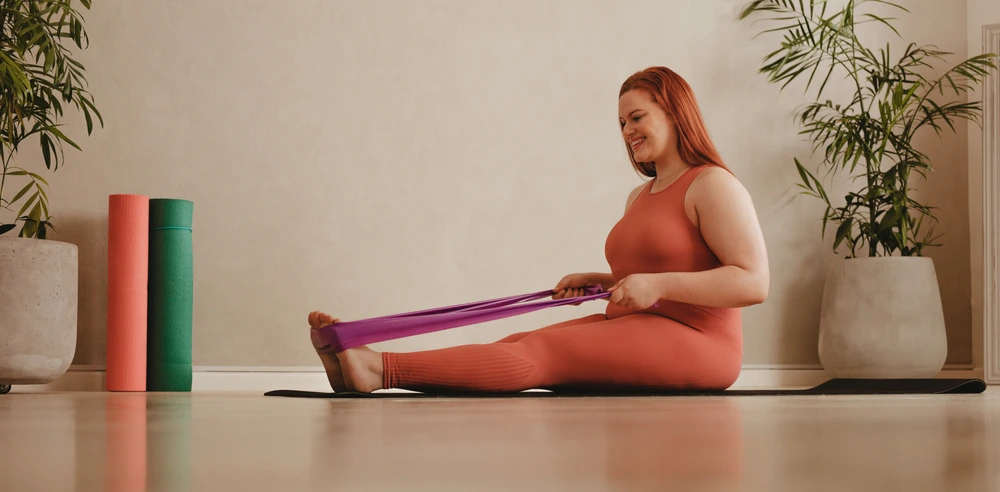 Frau in Sportkleidung trainiert mit einem Fitnessband im Sitzen, um gezielt an den Beinen abnehmen zu können.