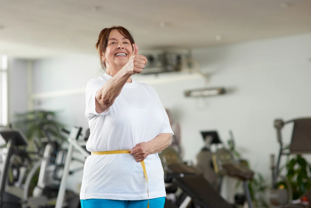 Ältere Frau misst ihre Taille im Fitnessstudio und zeigt zufrieden den Daumen hoch – Motivation und Erfolg beim Abnehmen im Alter.