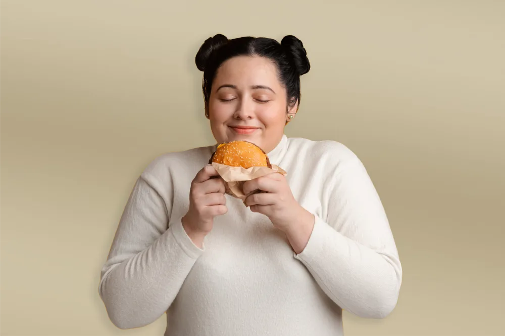 Junge Frau hält genüsslich einen Burger in den Händen und schließt die Augen vor Freude – ein typisches Bild für starken Heißhunger auf ungesunde Lebensmittel.