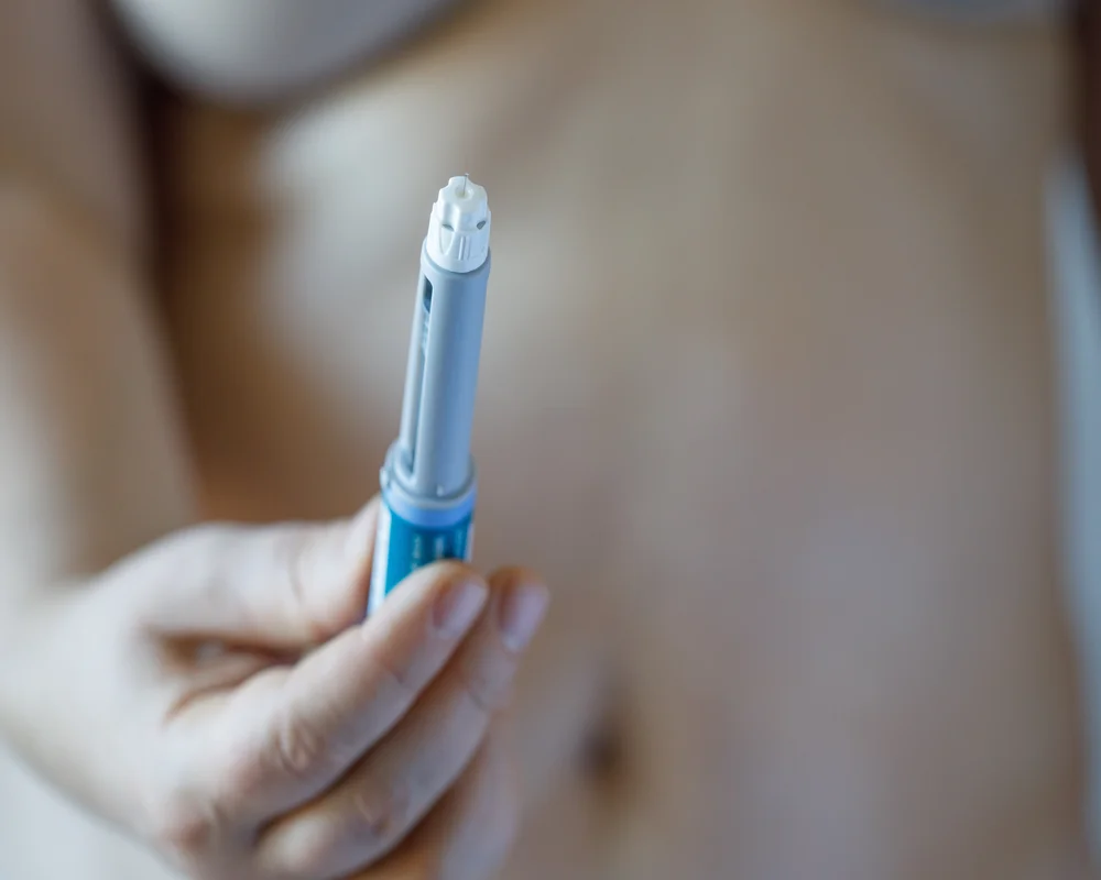 Eine Person hält eine Injektionshilfe in der Hand, die oft als Diabetes-Spritze zum Abnehmen verwendet wird und direkt am Bauch angewendet werden kann.