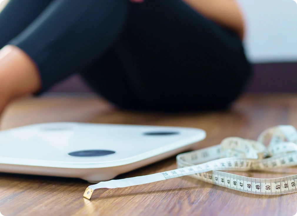 Nahaufnahme eines Maßbands und einer Waage auf dem Boden, während im Hintergrund eine Person sitzt – symbolische Darstellung dafür, wie ein BMI Rechner beim Einschätzen des eigenen Körpergewichts hilft.