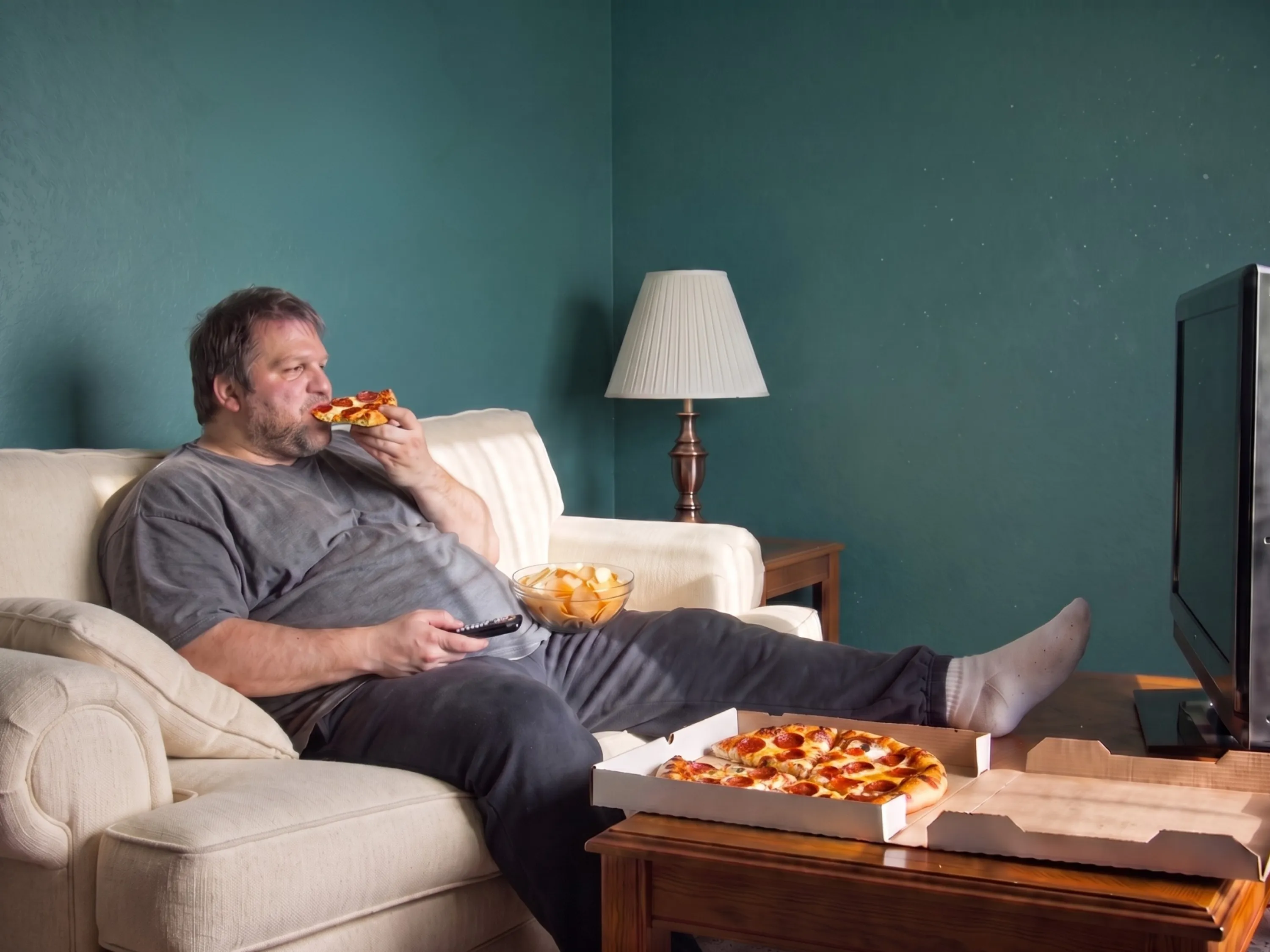 Mann sitzt vor dem Fernseher auf dem Sofa und isst ein Stück Pizza, daneben Chips und die restliche Pizza auf dem Tisch als Symbolbild für Essen aus Langeweile.