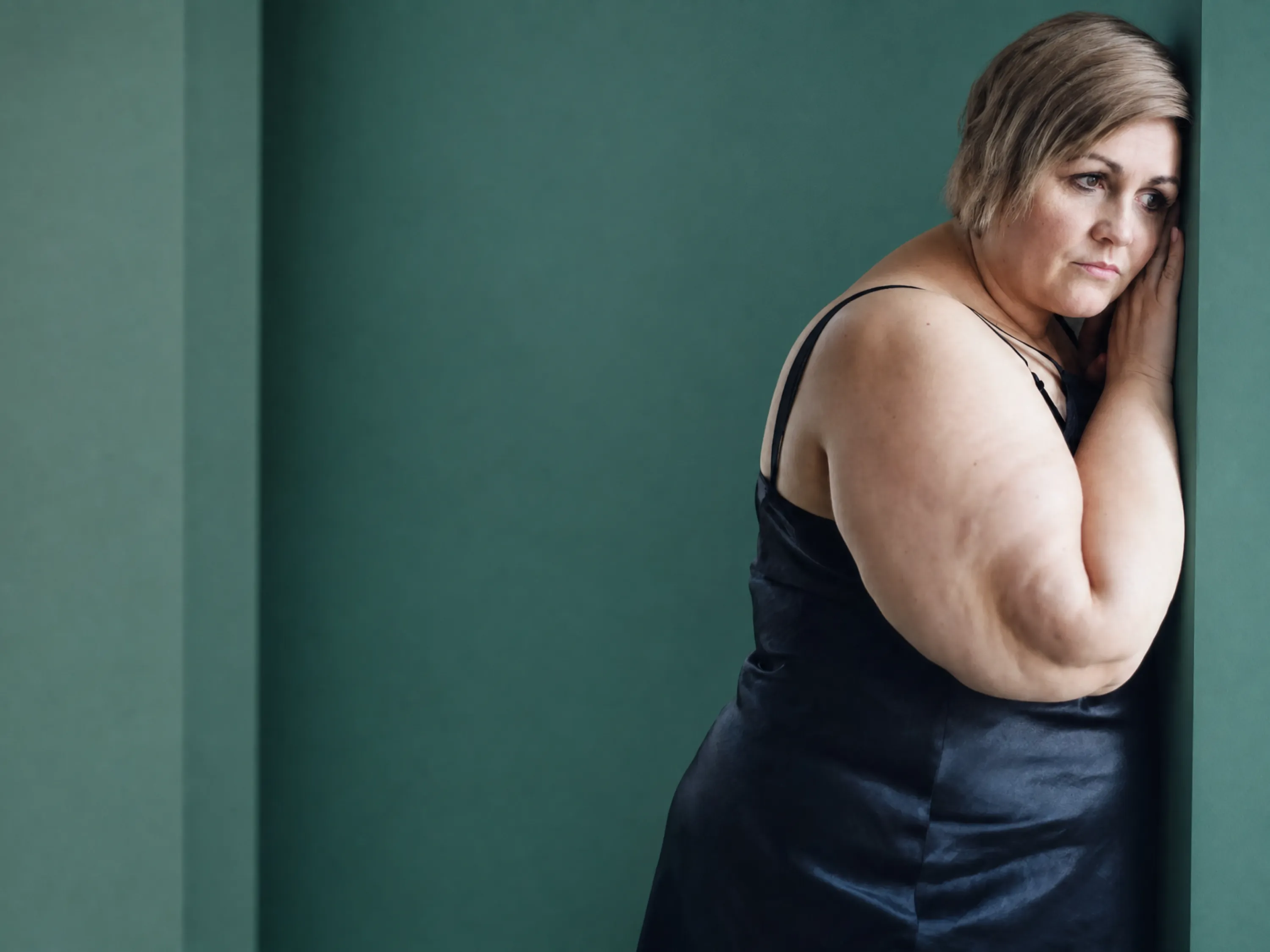 Nachdenkliche übergewichtige Frau lehnt traurig an einer Wand, was die emotionalen Folgen von Bodyshaming zeigt.