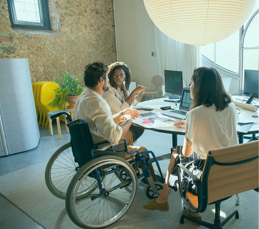 Three colleagues in a modern office having a discussion around a round table, one of them in a wheelchair, with laptops and documents on the table.