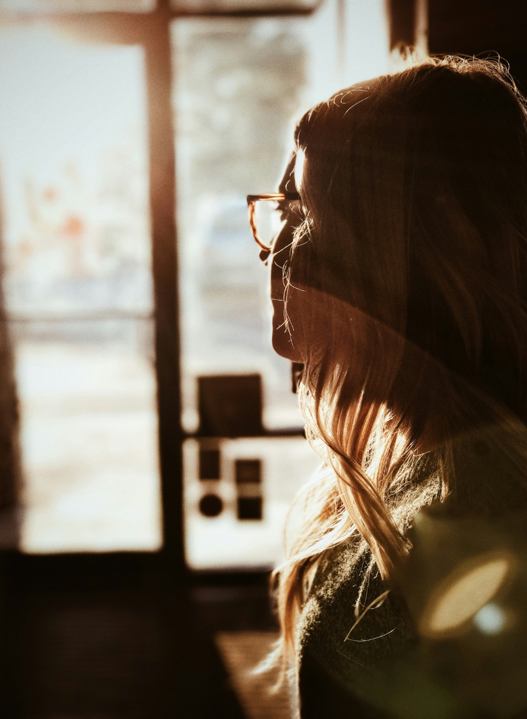 Person with glasses looking out window, backlit by warm sunlight