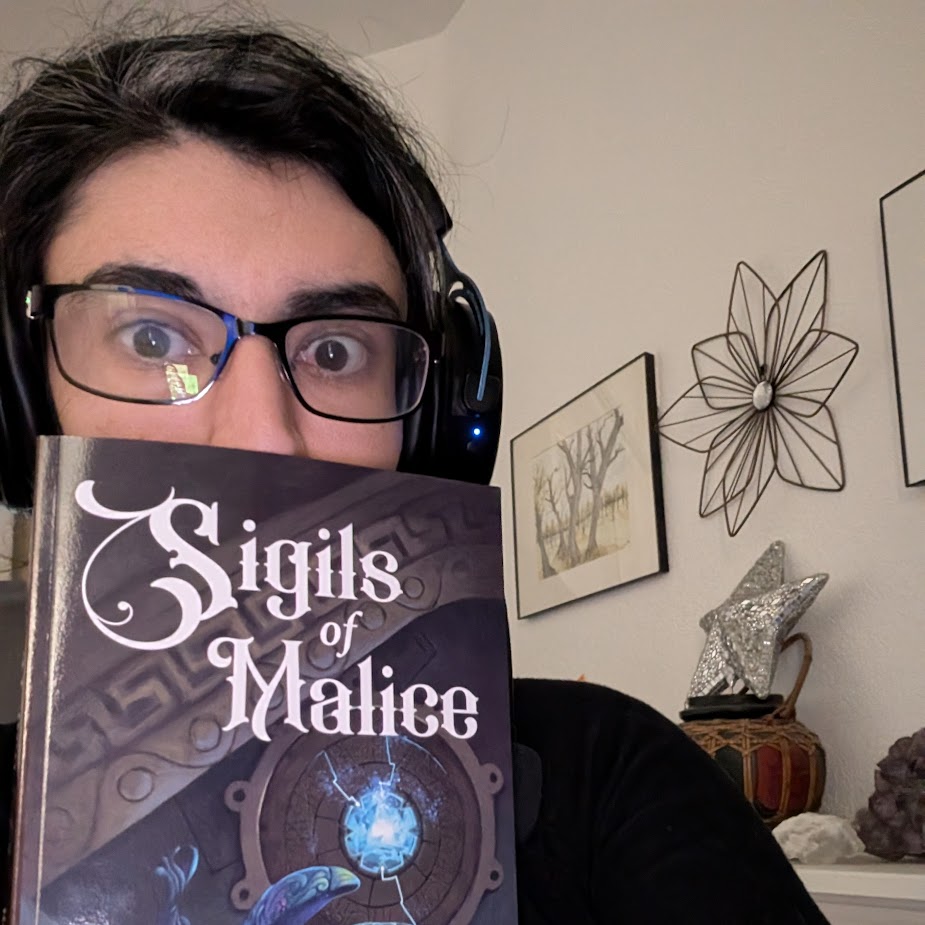 Picture of Rebecca Reyes without her glasses, smiling in front of two of her books on a decorated shelf. Sigils of Malice has a magical blue beetle before a glittering gem. Sigils of Blight is of a lantern in a jungle with burning ruins. There’s a stuffed unicorn and vines on the decorated shelf.
