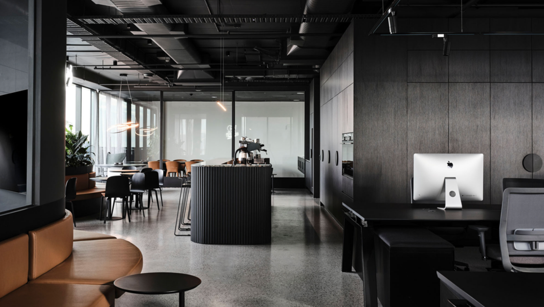 Modern office lounge with curved brown leather seating, black chairs, a central black coffee bar, and a desk with an iMac computer.