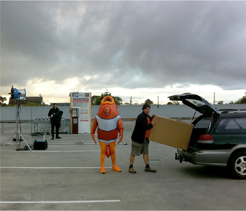 Person in an orange fish costume standing beside a man loading a large cardboard box into the back of a green station wagon in a parking lot under cloudy sky.