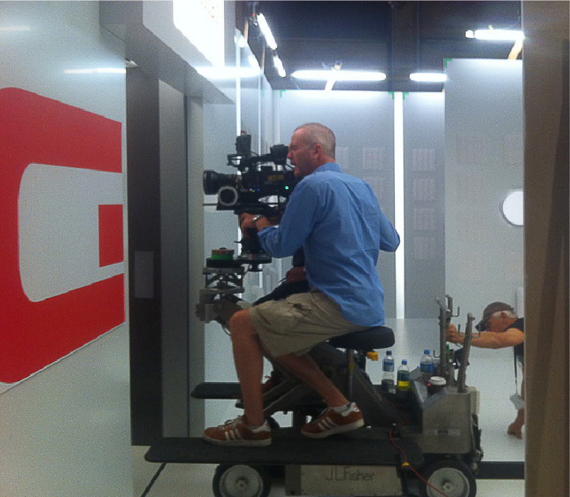 Man in a blue shirt and shorts operating a professional video camera mounted on a wheeled camera dolly inside a studio.