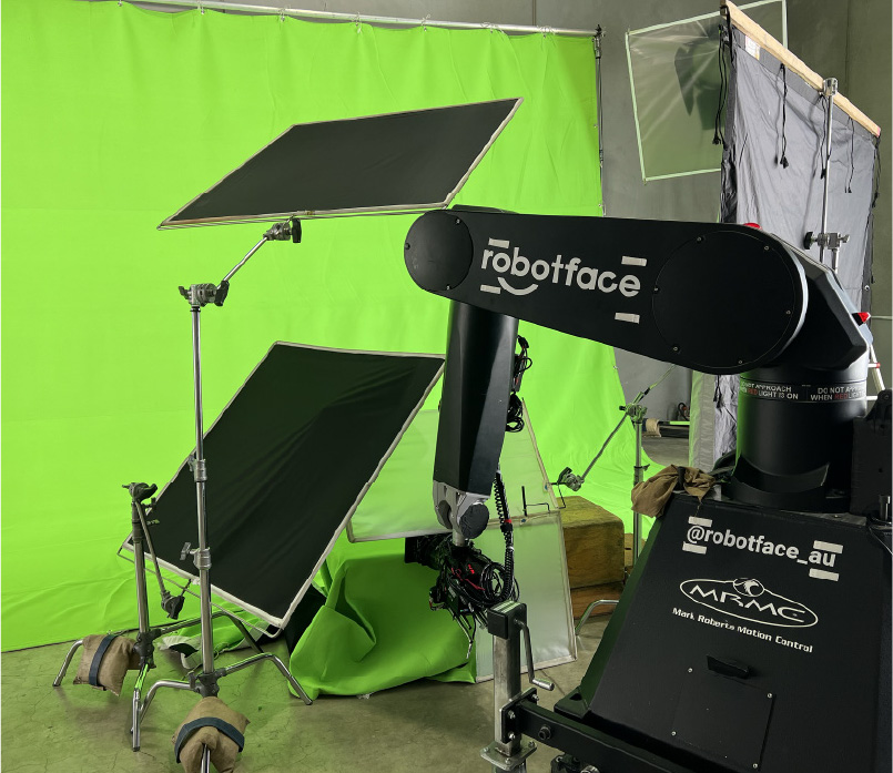 Robot arm labeled 'robotface' positioned in front of green screen with lighting equipment stands around it.