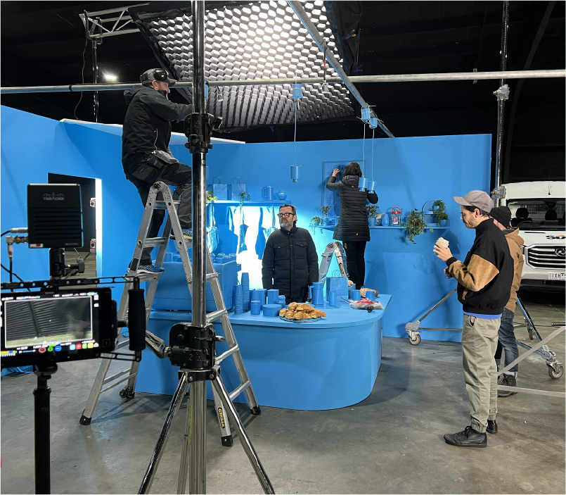 Film crew setting up a blue-themed indoor set with lighting equipment and crew members adjusting decorations and monitors.