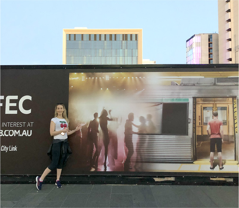 Woman in casual clothing posing and pointing at an advertising billboard showing silhouettes of people dancing and a man standing in a train doorway.