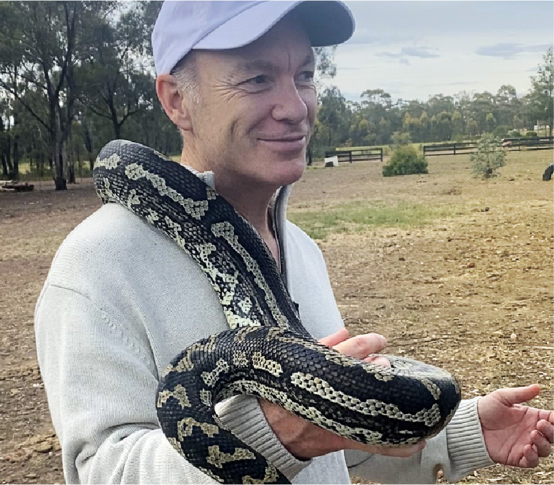Man wearing a light gray cap and sweater holding a large black and beige patterned snake outdoors in a rural area.