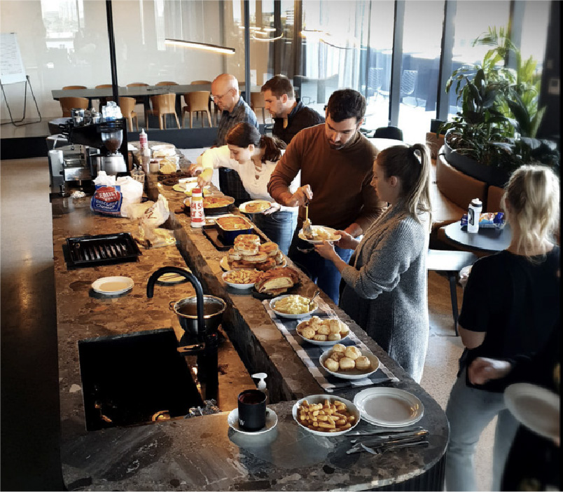 Group of people serving themselves food from a buffet table in a modern office kitchen with various dishes and snacks laid out.