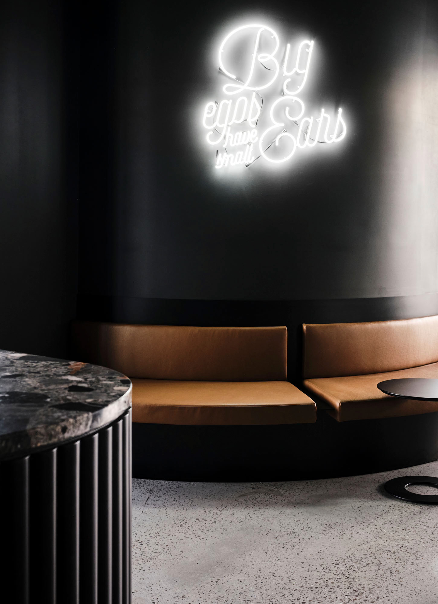 Modern seating area with brown leather benches against a black wall illuminated by a neon sign reading 'Big ears have small ears'.