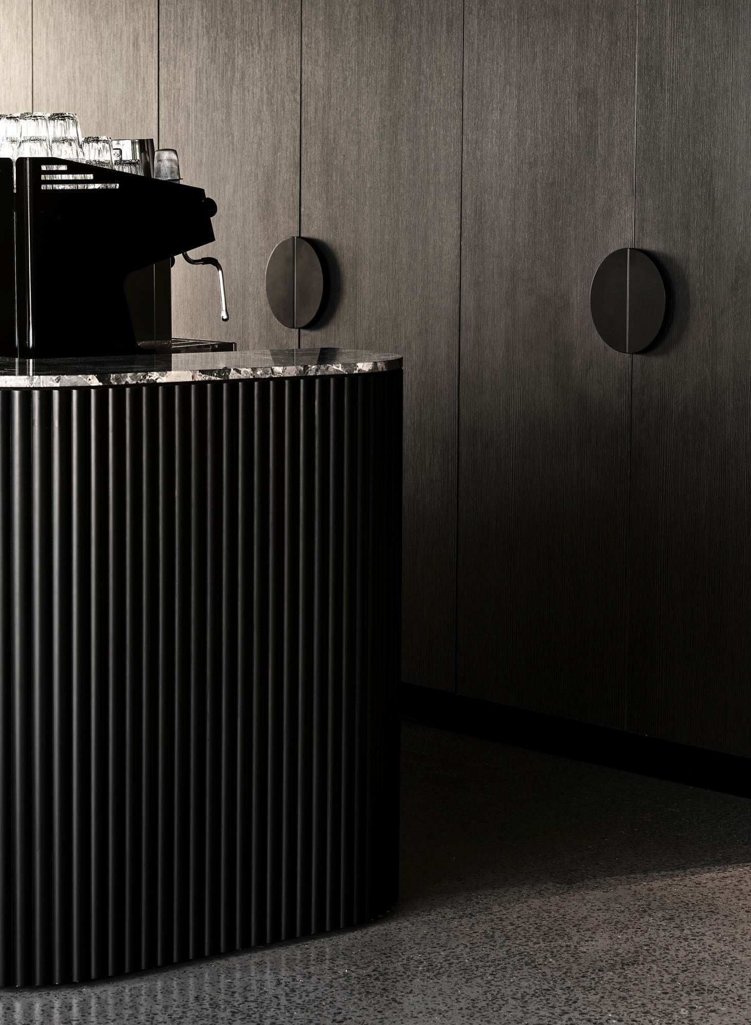 Modern black coffee machine on a ribbed black counter with a dark textured wall and circular cabinet handles in the background.