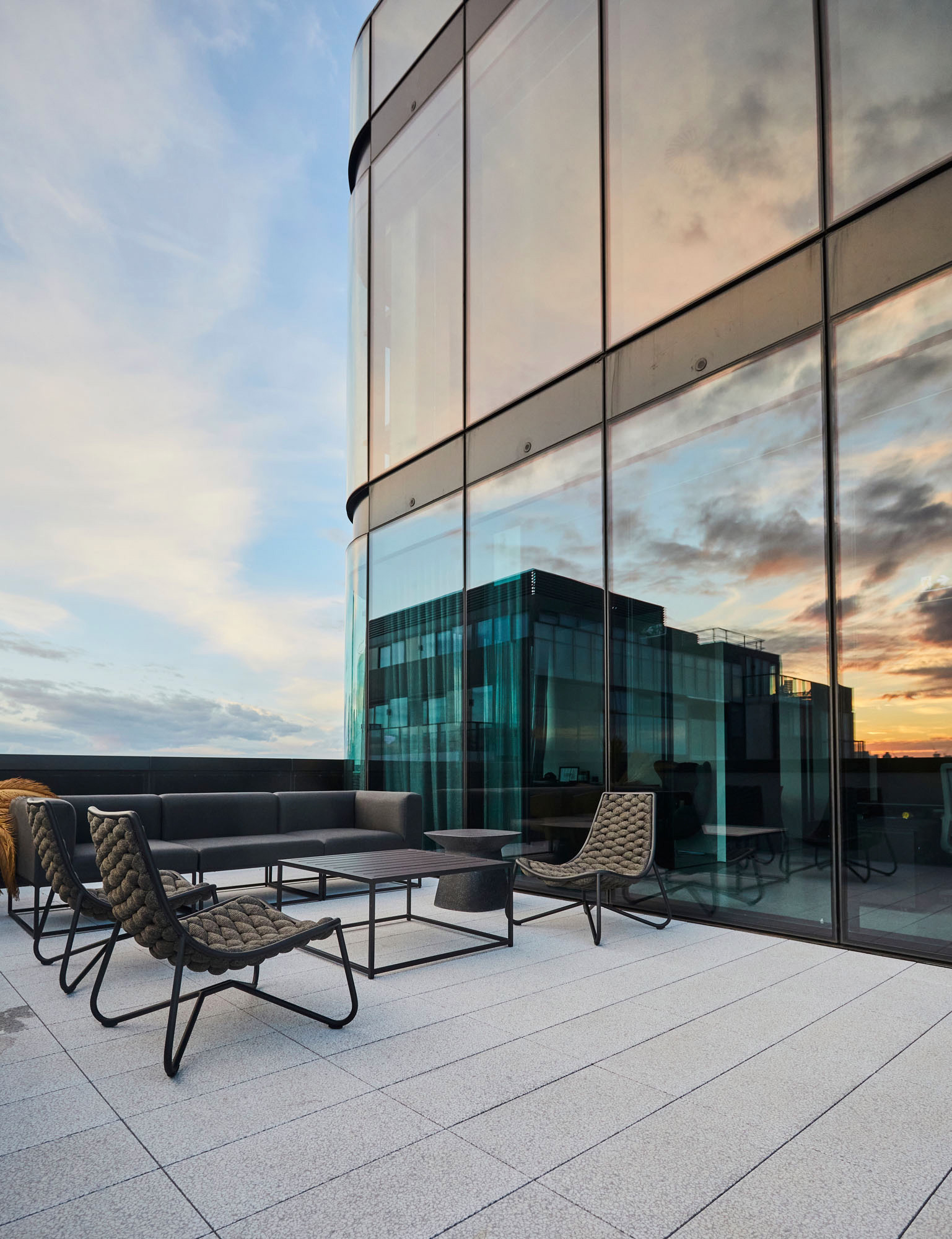 Modern outdoor patio with gray cushioned chairs and sofa on tiled floor next to a glass building reflecting a sunset sky.