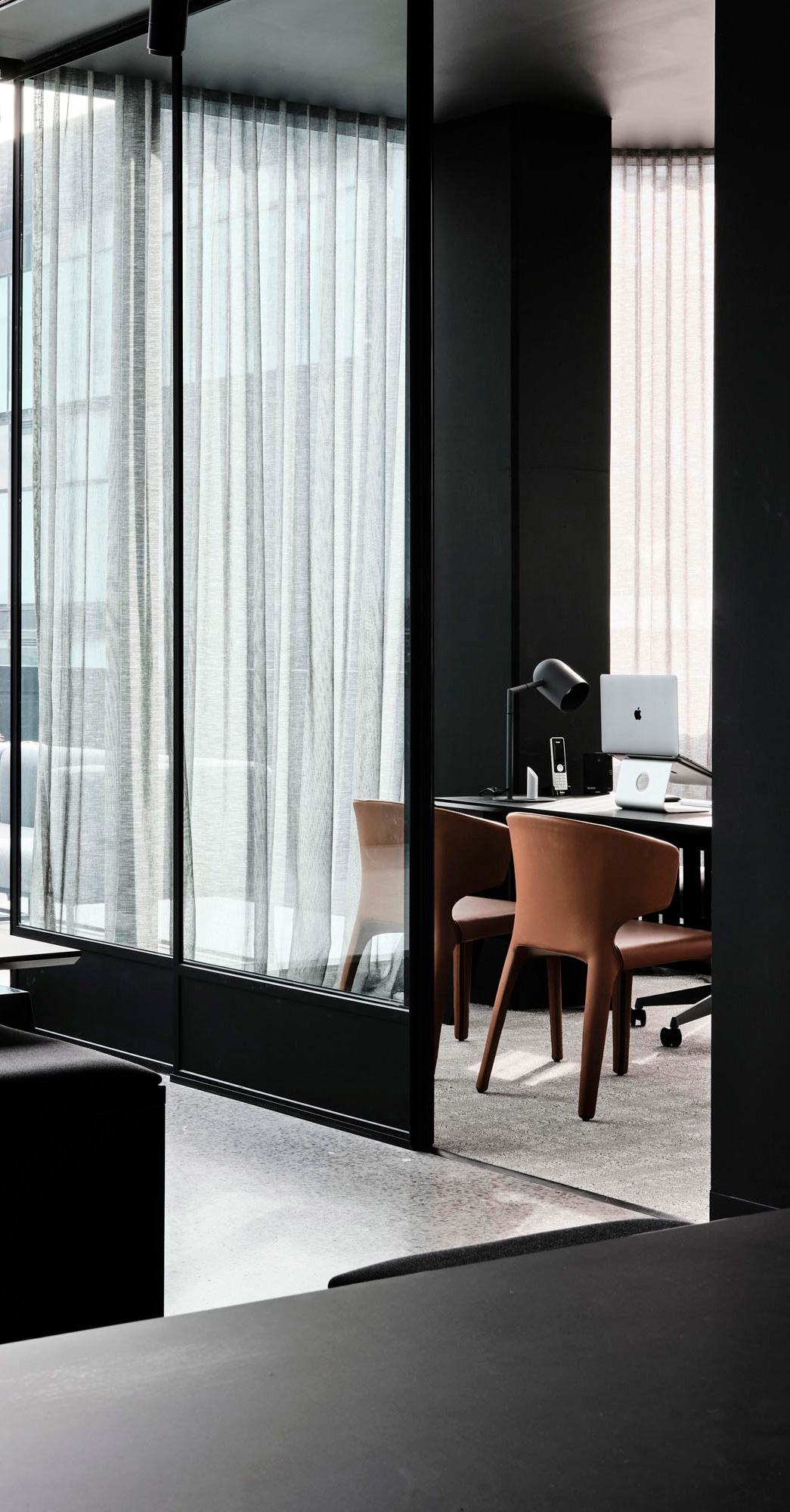 Modern office room with glass walls, brown chairs, a desk lamp, and an Apple laptop.