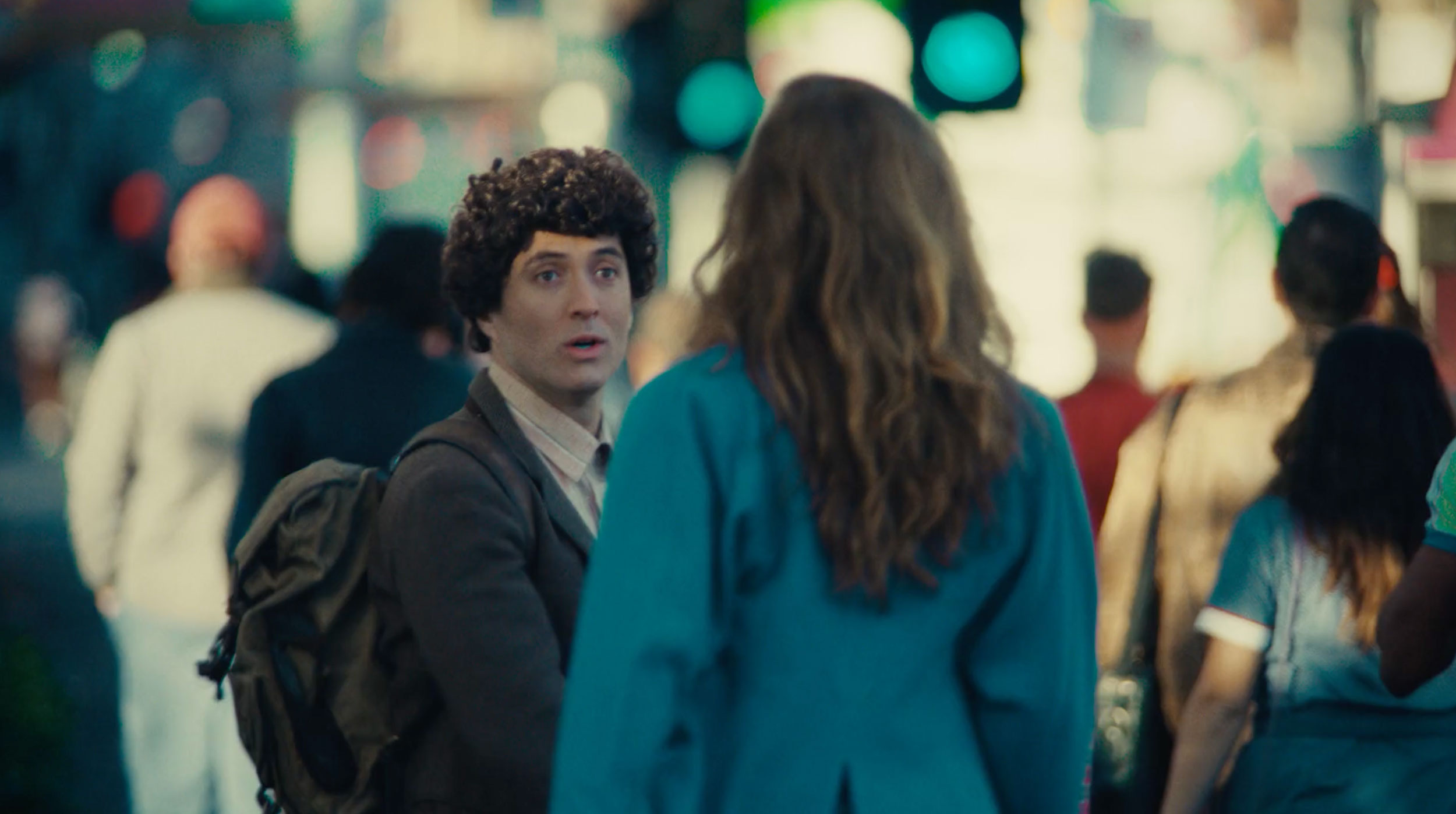 Man with curly hair and backpack looks surprised while talking to a woman with long brown hair on a busy city street.