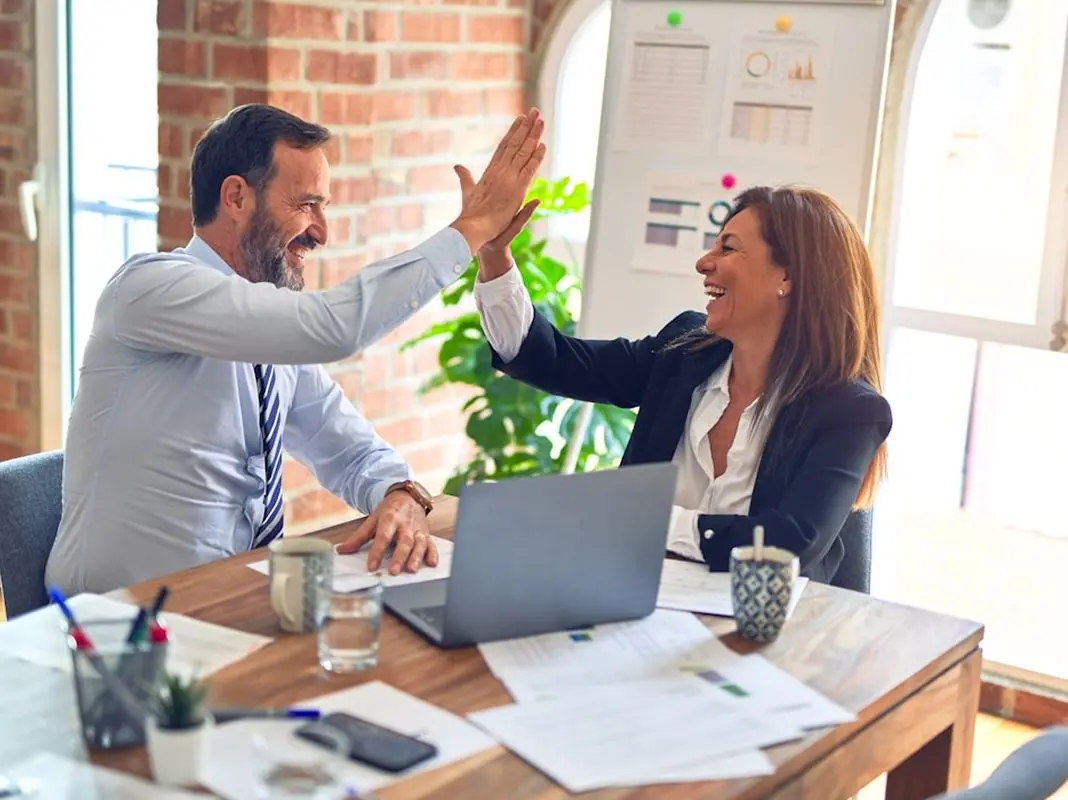 Two business colleagues smiling and giving a high five across a desk with a laptop and documents.