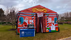 a vibrant scene set in a park. Dominating the center of the image is a large, red tent with a whimsical Christmas-themed design. The tent is adorned with a sign that reads "SANDBOX TRAINING WORKSHOP? Help Wanted! High-quality instructors needed. Contact us for more information." The tent is situated on a grassy area, with a solitary tree standing in the background. A blue table is also present in the scene, bearing a sign that reads "FREEDOFUND.COM".