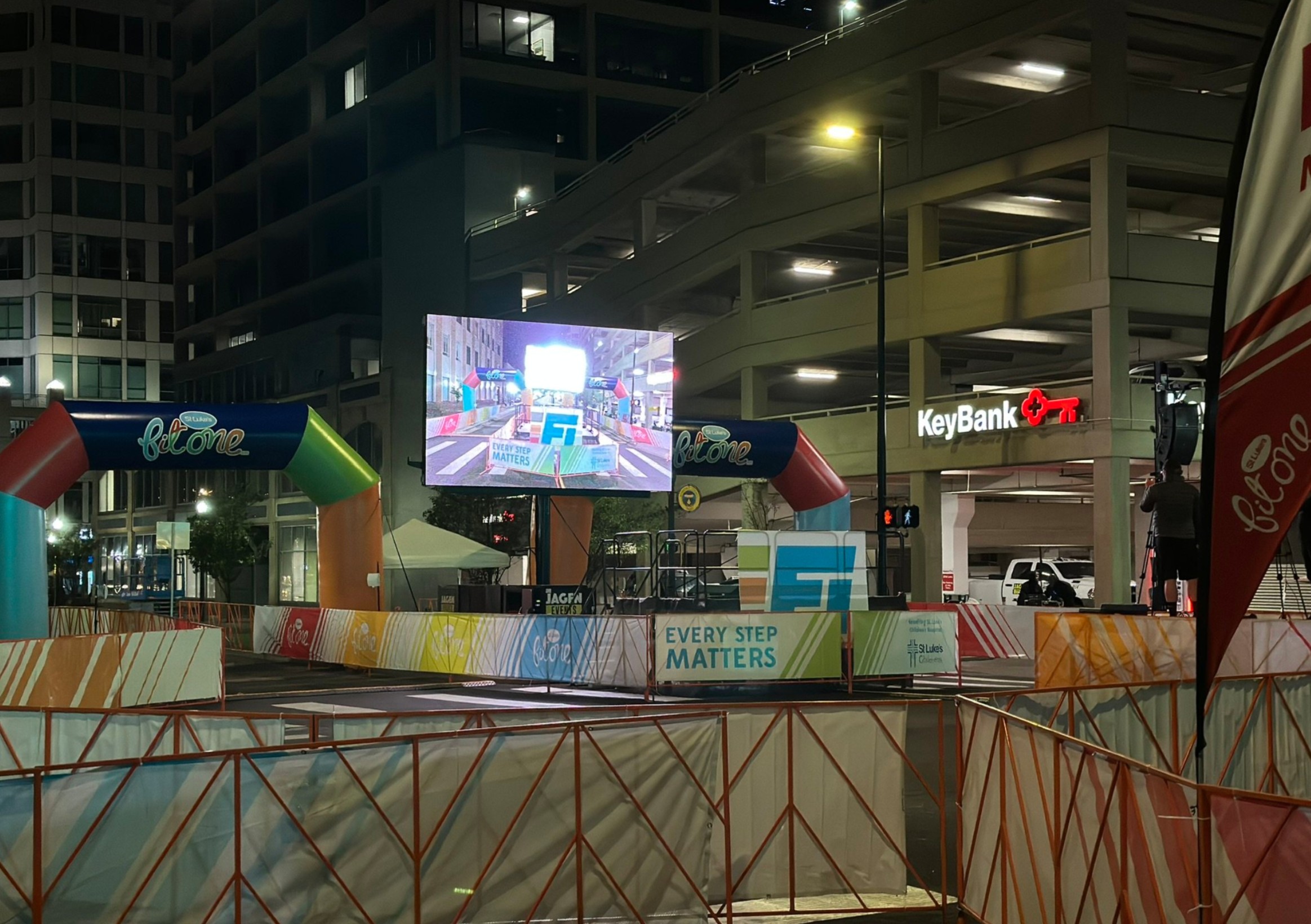 Mobile LED screen broadcasting live race coverage at a nighttime urban fitness event, positioned near a colorful start/finish arch and KeyBank garage, enhancing visibility for participants and spectators.