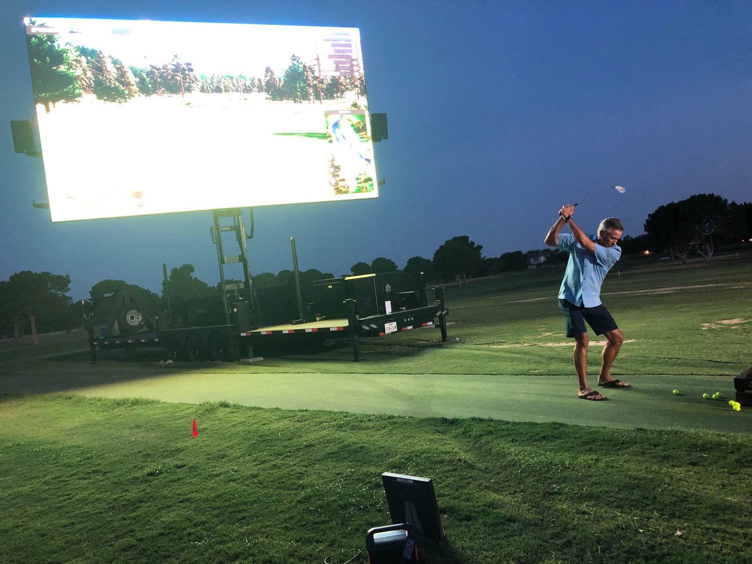 Man swinging a golf club at dusk in front of a large mobile LED screen displaying a virtual golf course on a driving range. Perfect for outdoor sports events and interactive digital experiences.