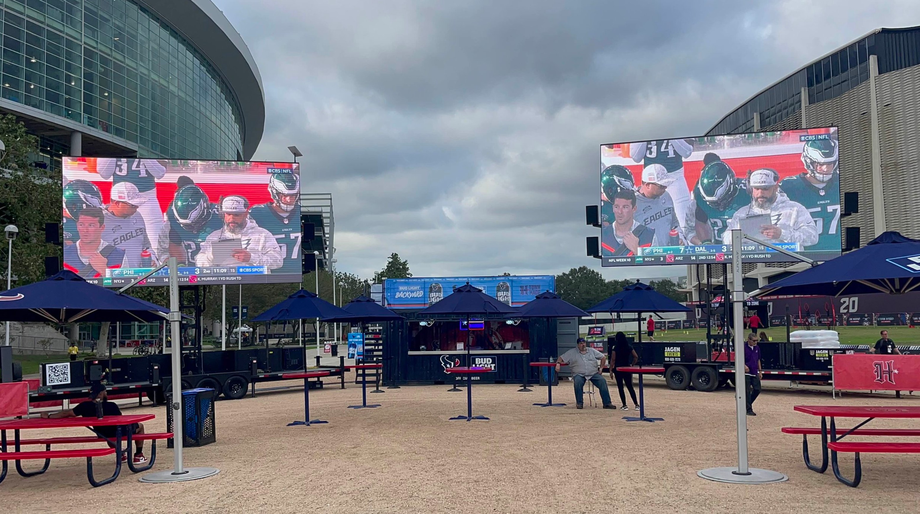 Two large LED screens streaming a live football game outside a stadium at a Bud Light fan zone, with picnic tables, umbrellas, and a branded bar setup for public viewing.