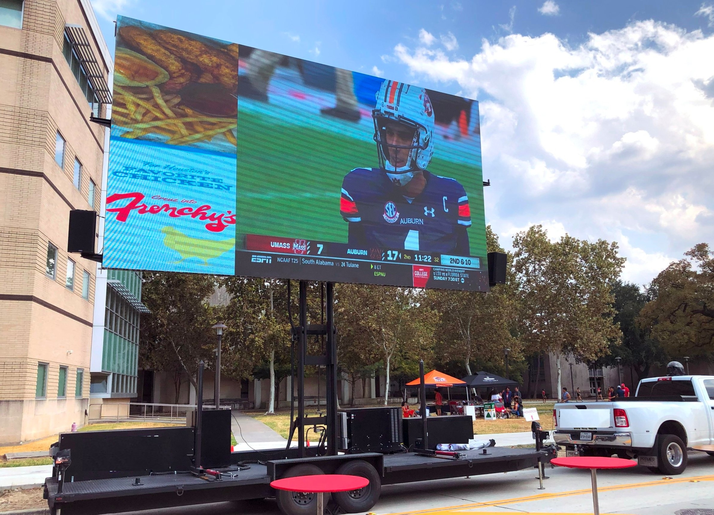 Large mobile LED screen streaming a live college football game and advertisement at an outdoor campus tailgate event, mounted on a trailer with full audio support and promotional branding.