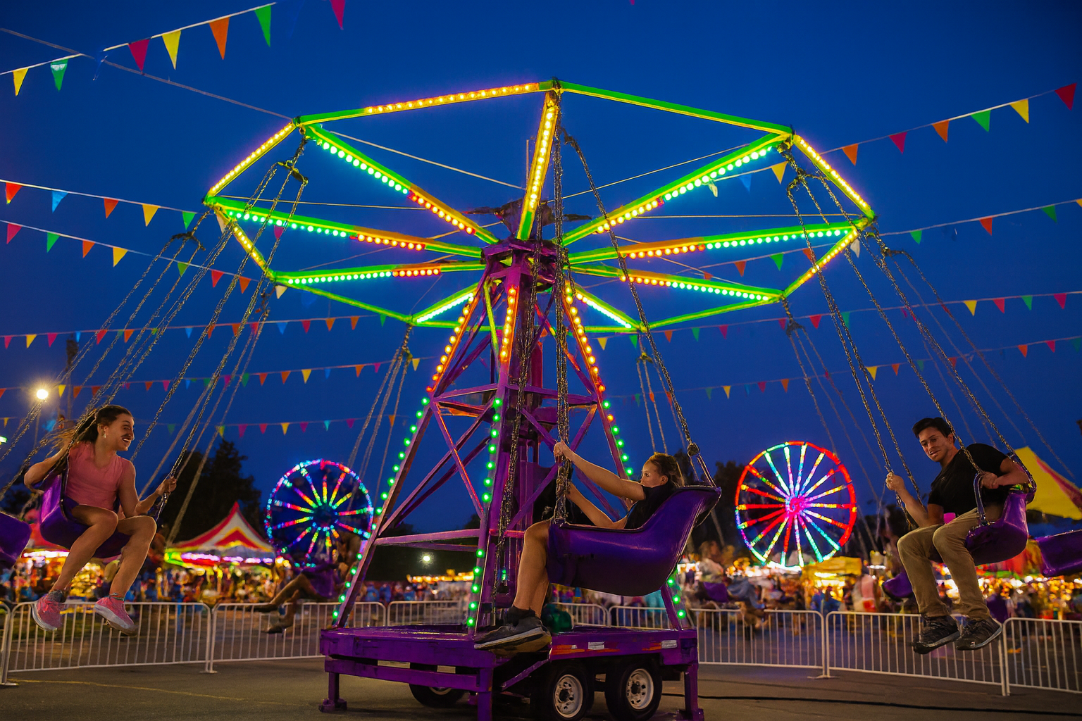Three carnival staff members in red Freedom Fun shirts ride a colorful spinning carnival ride with arms raised and smiling faces. The ride features bright red, yellow, green, and orange bucket seats against a vibrant backdrop of tents, flags, and a Ferris wheel at a busy outdoor fair.