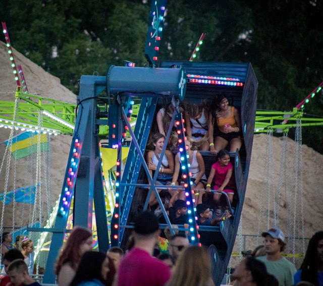 A carnival ride called Phantom’s Revenge swings a group of smiling teens through the air under colorful LED lights at night. The ride structure is illuminated with blue and red lights, surrounded by crowds and other glowing attractions in the background.