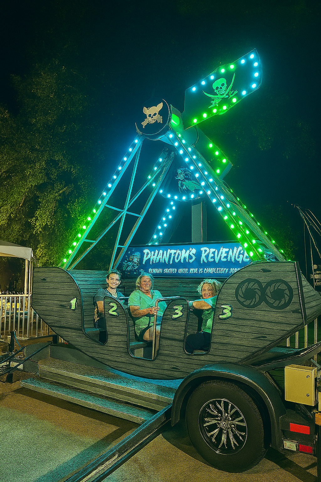 Phantom’s Revenge carnival ride stands on a grassy field under a clear blue sky, empty and ready for action. The pirate ship-themed ride is lit up with bright green and blue LED lights along the frame and flag, with four numbered seat sections visible on the wooden-style body. A tree line stretches across the horizon in the distance.