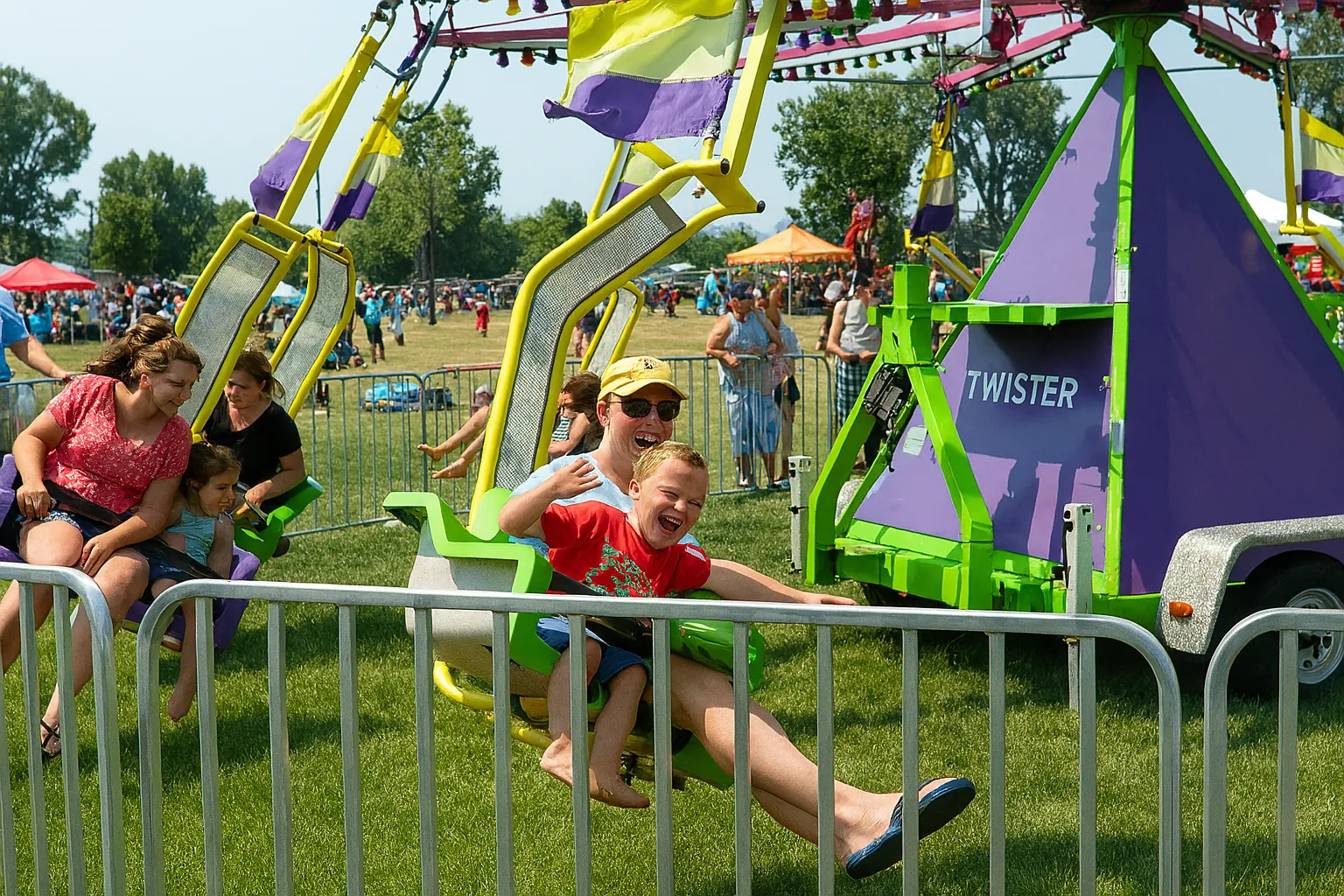 A joyful family rides the Twister swing carnival ride at an outdoor festival. A smiling child sits in front of a parent on a green and white swing seat, laughing with excitement. Other families are also enjoying the ride, with colorful flags above and crowds and tents visible in the sunny background. The purple and green triangular ride base labeled “TWISTER” stands out in the center.