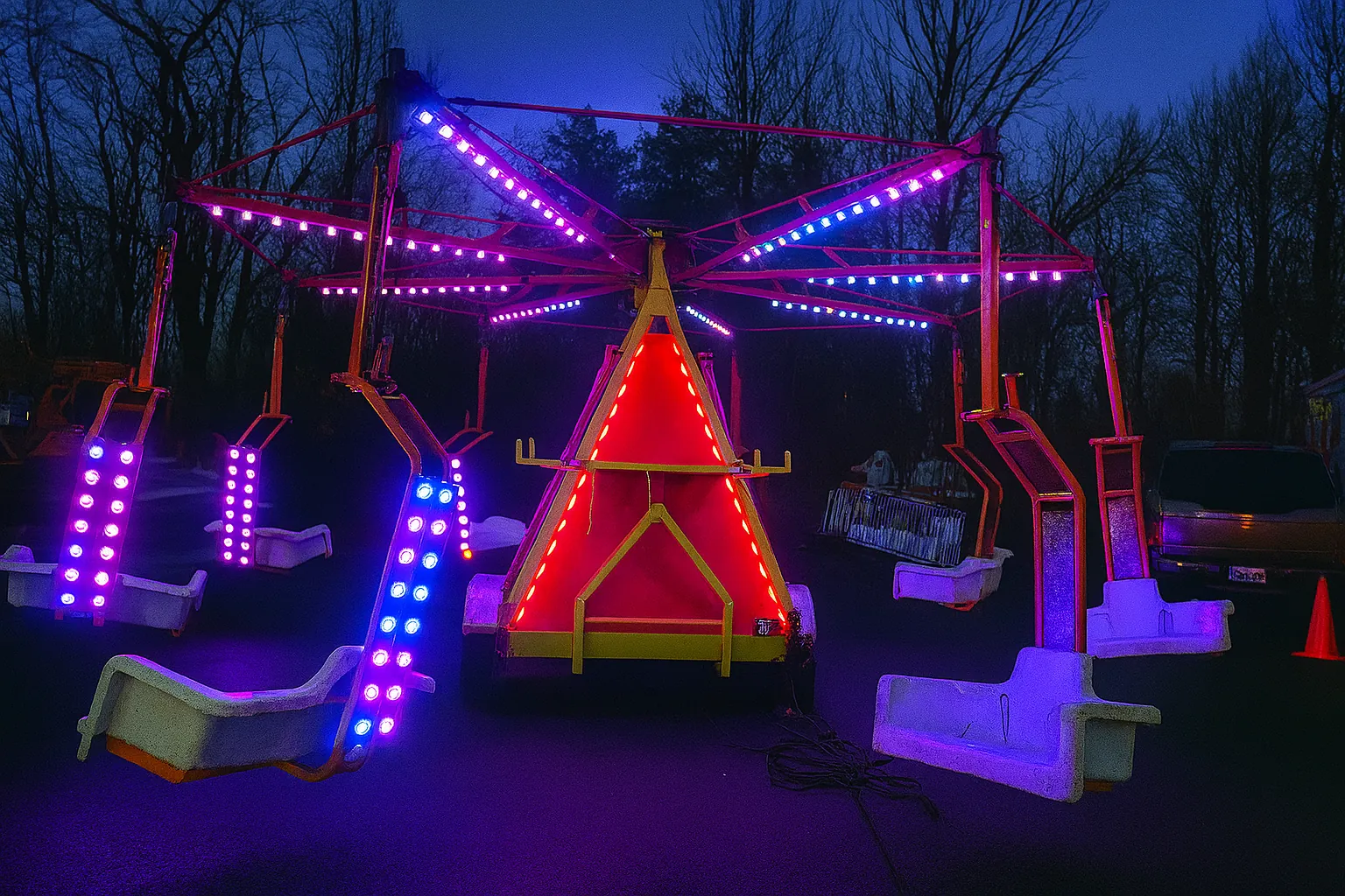 A brightly lit carnival swing ride glows in the evening darkness, featuring vibrant LED lights in pink, blue, and red. The white swing seats are suspended from a central triangular base, which is outlined with red lighting. The background shows leafless trees and a dimly lit area, creating a striking contrast with the ride’s colorful illumination.