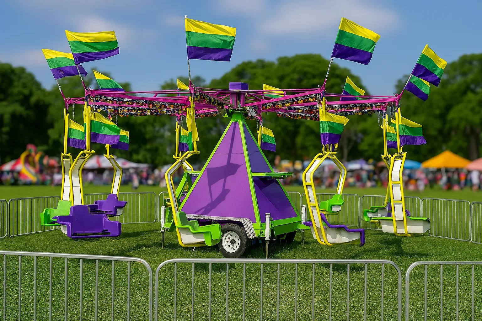 A stationary Twister swing carnival ride stands on a grassy field, surrounded by metal fencing. The ride features a bold purple and green triangular center with multiple yellow, green, and purple flags atop each swing arm. The background includes a cloudy sky and industrial buildings, suggesting setup before an event.