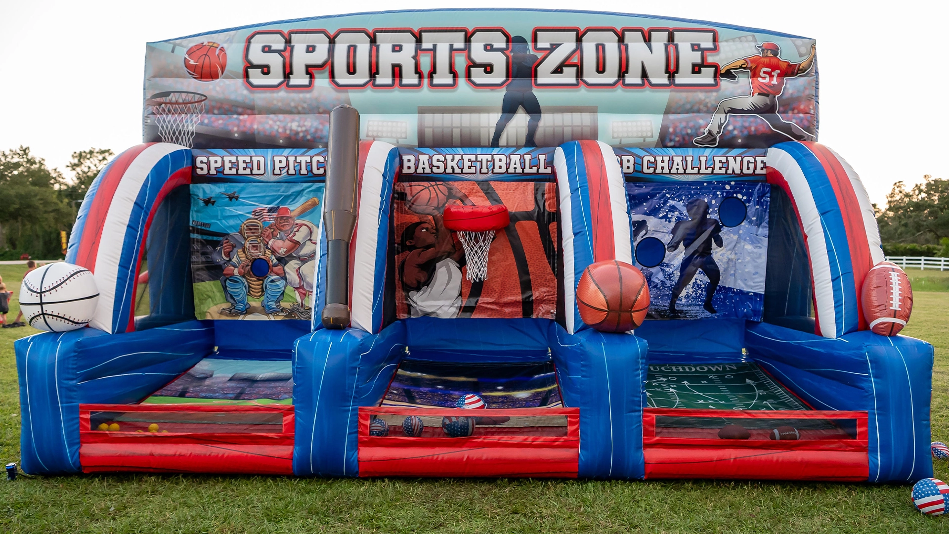 In a vibrant park, a group of six people are gathered around a large inflatable obstacle course. The course is a striking combination of red and blue, with a large inflatable football and baseball on the left side, and a large inflatable soccer goal on the right side. The people are engaged in various activities, some standing and others in motion, suggesting a sense of camaraderie and fun. The park is lush with green grass, and in the distance, trees can be seen.
