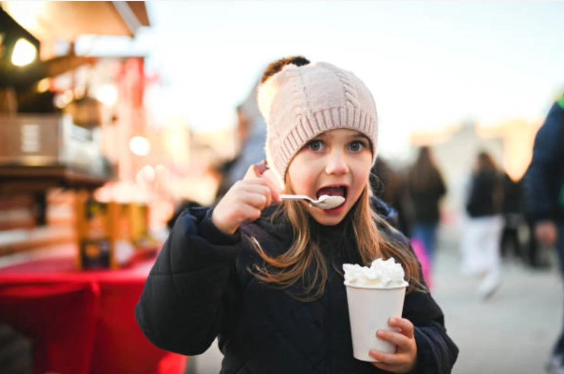 Young girl in winter hat happily drinking hot cocoa at outdoor holiday market.
